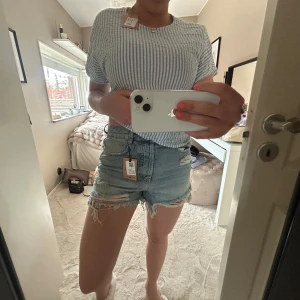 Ljusblå jeansshorts med slitningar - Säljer ett par ljusblå jeansshorts med råa kanter och slitna detaljer framtill. Shortsen har klassisk femficksdesign, knapp  och de är Perfekta för varma dagar.