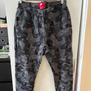 Nike camo mjukisbyxor med snörning - Säljer ett par grå och svarta mjukisbyxor från Nike med camouflagemönster. Byxorna har snörning i midjan, muddar vid bensluten och är tillverkade i en mjuk bomulls- och polyestermix. Perfekta för en avslappnad stil.