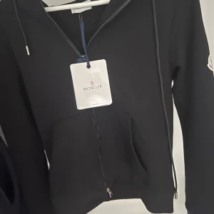 Moncler kofta - Säljer en svart hoodie från Moncler med dragkedja framtill och fickor. Tröjan har huva med röd, vit och blå detalj samt Moncler-logga på ärmen. Perfekt för en stilren och sportig look. Finns även kvar kvitto på den.