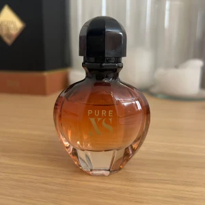 Pure XS parfym från Paco Rabanne - En elegant parfymflaska från Paco Rabanne, Pure XS. Flaskan är rundad med en varm bärnstensfärgad ton som övergår till klar glas nedtill och har ett svart lock. Perfekt för dig som gillar lyxiga och sensuella dofter.