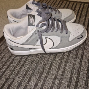 Nike Dunk Low i grått och vitt - Säljer ett par Nike Dunk Low sneakers i grått och vitt med klassisk swoosh på sidan. Skorna har snörning, rund tå och platt sula. Perfekta för dig som gillar stilrena och sportiga sneakers.