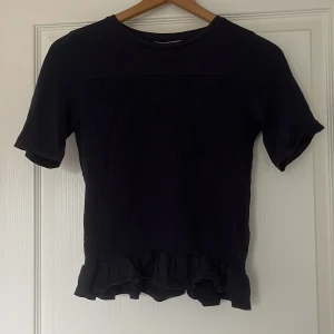 Mörkblå med volangkant - Säljer en mörkblå topp med korta ärmar och volangdetalj längst ner. Toppen har en lätt rynkad struktur på tyget som ger en snygg effekt. 