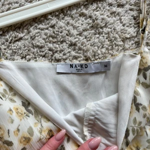 Blommig klänning från NA-KD - Säljer en romantisk blommig klänning från NA-KD i storlek 36. Klänningen har smala axelband, v-ringning och volanger. Mönstret är små gula och gröna blommor på vit botten, perfekt för varma dagar.