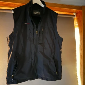 Fransk vindväst - Svart väst med hög krage och dragkedja framtill samt två fickor med dragkedja. Västen har en diskret detalj i form av en liten färgad linje på bröstet. Perfekt för lager-på-lager och enkel att matcha.