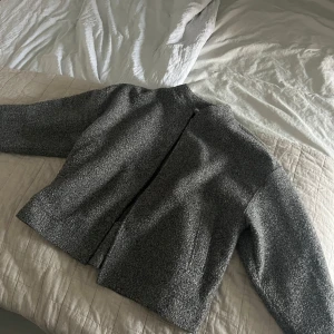 Grå ullkappa med minimalistisk design - Säljer en stilren grå kappa i ullblandning med rak passform och diskreta knappar. Jackan har lång ärm och en klassisk, enkel look som passar perfekt till höst och vinter. Perfekt för dig som gillar en clean och modern stil.