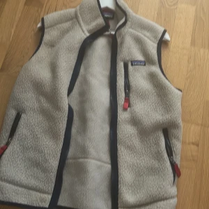 Beige fleeceväst från Patagonia - Säljer en beige fleeceväst från Patagonia med svarta detaljer och dragkedjor med röda dragare. Västen har hög krage och två sidofickor samt en bröstficka med dragkedja. Perfekt för lager-på-lager och outdoor.