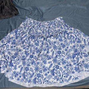 Vit och blå blommig kjol med volang - Supersöt kortkjol med blått blommönster på vit botten. Kjolen har volanger och en smockad midja med knytband. Perfekt för en fräsch och somrig look!