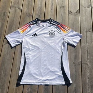 Tysklands fotbollströja från Adidas - Säljer en vit Tyskland fotbollströja från Adidas med svarta detaljer och ränder i rött, gult och svart på axlarna. Broderad landslagslogga med fyra stjärnor på bröstet. Kortärmad modell i lätt och ventilerande material. Säsong 23/24.