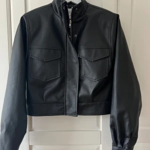 Svart skinnjacka med dragkedja - Säljer en svart skinnjacka i kortare modell med hög krage och dragkedja framtill. Jackan har två stora bröstfickor med lock och långa ärmar. Jackan är från NA-KD Josefine HJs kollektion, storlek 32 passar xs/s skulle jag säga. Använd en gång