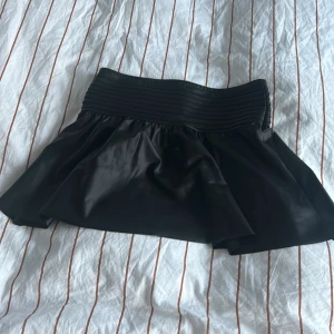 Svart kortkjol från Gina Tricot - Säljer en svart kortkjol från Gina Tricot med bred resår i midjan. Kjolen har en glansig finish och är lätt utställd för en snygg siluett. Perfekt att styla med en enkel topp eller hoodie.