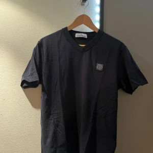 Mörk blå t-shirt från Stone Island - Mörk blå t-shirt från Stone Island med v-ringad hals och klassisk logotyp-patch på bröstet. Tillverkad i mjuk bomull och har korta ärmar. Perfekt för en stilren och avslappnad look. Nu pris på tröjan är 1 699kr 