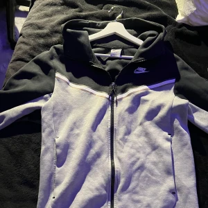 Nike tech  ziphoodie i svart och grå  - Storlek xs men passar även s. Snygg ziphoodie från Nike i svart och ljusgrått med vit logga på bröstet. Jackan har huva, dragkedja och två fickor framtill. Perfekt för en sportig och avslappnad stil.
