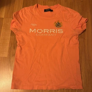 Persikofärgad t-shirt från Morris - S - Säljer en persikofärgad t-shirt från Morris med tryckt logga och emblem på bröstet. T-shirten har rund halsringning och korta ärmar. Perfekt för en avslappnad och stilren look.