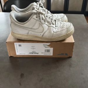 Vita Nike Air Force 1 sneakers - Klassiska vita Nike Air Force 1 sneakers med snörning och rund tå. Skorna har en platt sula och är tillverkade i skinn med perforerade detaljer framtill för extra ventilation. Perfekta för dig som gillar en tidlös och sportig stil.