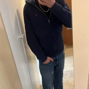 Mörkblå hoodie från Polo Ralph Lauren - Säljer en mörkblå hoodie från Polo Ralph Lauren med dragkedja och huva. Tröjan har två fickor framtill och den klassiska röda loggan broderad på bröstet. Perfekt för en avslappnad stil.