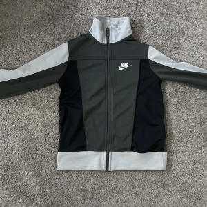 Nike träningsjacka i svart och grått - Sportig Nike jacka i svart och grått med dragkedja framtill och hög krage. Jackan har vita detaljer och klassisk Nike-logga på bröstet. Perfekt för träning eller som en snygg vardagsjacka.