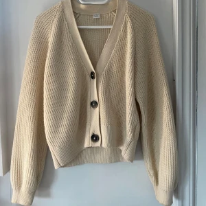 Beige stickad kofta med knappar - Säljer en beige stickad kofta med stora svarta knappar framtill. Koftan har lång ärm, ribbade muddar och en avslappnad passform. Perfekt att slänga på sig när det är kyligt ute.