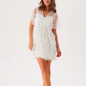 Vit blommig kortklänning med volang och v-ringning,3d blommor  - Superfin vit kortklänning med v-ringning och transparenta ärmar. Klänningen har blommigt mönster i 3D, volangdetaljer och en luftig passform. Perfekt för sommarens alla tillfällen! Har endast använts en gång, inga skavanker! Som ny!