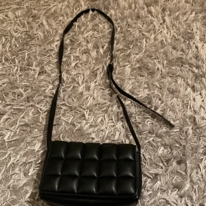 Svart quiltad axelväska - Säljer en svart quiltad axelväska med lång justerbar rem. Väskan har en stilren och modern design med rutmönstrad vaddering på framsidan. Perfekt för att bära det viktigaste med stil.