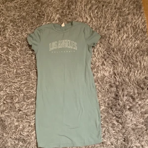 Grön t-shirtklänning med tryck 'Los Angeles' - Säljer en ljusgrön t-shirtklänning med texten 'Los Angeles California' framtill. Klänningen har korta ärmar och rak passform, perfekt för en avslappnad stil. Mjuk och skön bomullskänsla.