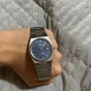Tissot PRX Powermatic 80 med blå urtavla - Säljer en stilren Tissot PRX Powermatic 80 herrklocka med blå urtavla och datumvisning. Klockan har ett silverfärgat armband i rostfritt stål och en transparent baksida som visar urverket. Perfekt för den som söker en elegant och tidlös accessoar. Funkar som den ska låset är lite defekt men syns inte alls. Banden kan bytas enkelt också ifall man verkligen vill byta den. Billig för en anledning och ja det är det du tror...