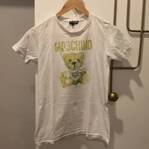 Vit t-shirt med nalletryck från Moschino - Vit t-shirt från Moschino med stort gult MOSCHINO-tryck och en nalle på framsidan. Klassisk passform och rund halsringning. Perfekt för dig som gillar statement-plagg och coola prints.