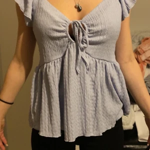 Ljuslila blus med knytning - Säljer en ljuslila blus med v-ringning och knytning framtill. Blusen har korta volangärmar och är tillverkad i ett lätt, krinklat material. Den har en lös passform nedtill och en feminin siluett.