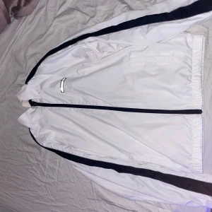 Vit vindjacka från Ellesse med svarta detaljer - Säljer en vit vindjacka från Ellesse med svarta ränder längs ärmarna och dragkedja framtill. Jackan har en enkel och sportig design med Ellesse-logga på bröstet. Perfekt för dig som gillar streetwear och sportig stil.