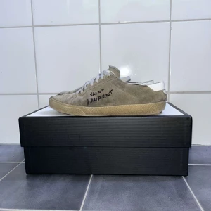 Saint Laurent  - Säljer ett par beigea Saint Laurent sneakers i mocka med vit sula och vita skosnören. Bara skorna. Större skador finns i bild 4 och 5 finns hål.  Sidan har handskriven logga och skorna har en klassisk, låg profil med rund tå. Perfekta för dig som gillar stilrena och exklusiva sneakers. 