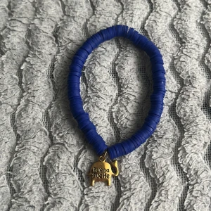 Blått armband med guldberlock - Snyggt armband med blåa platta pärlor och en liten guldberlock i form av en gris med texten 'Buena Suerte'. Perfekt accessoar för att addera färg till din outfit.