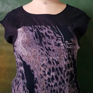 Svart topp med abstrakt mönster - Snygg svart topp med korta ärmar och ett abstrakt mönster i lila och grå toner framtill. Toppen har rund halsringning och är tillverkad i ett mjukt material som känns bekvämt mot huden. Perfekt för dig som vill ha något unikt i garderoben.
