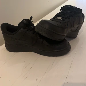 Svarta Nike Air Force 1 sneakers - Säljer ett par helsvarta Nike Air Force 1 sneakers med klassisk design. Skorna har snörning, rund tå och platt sula. Perfekta för dig som gillar stilrena och tidlösa sneakers med ikonisk Nike-logga på sidan och bak.