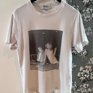 Vit t-shirt med tryck från Fiore Studios - Vit t-shirt från Fiore Studios i storlek XS med ett svartvitt tryck på framsidan som föreställer två personer vid ett bord. Klassisk passform och rund halsringning. Perfekt för dig som gillar stilrena plagg med coolt motiv. Og storlek är xs men den sitter mer som en xxs.