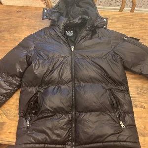 Svart pufferjacka  - Säljer en svart pufferjacka från LON med huva och dragkedja framtill. Jackan har två framfickor med dragkedja och en extra ficka på ärmen. Perfekt för kyliga dagar. Pris går att diskutera vid en snabb affär!