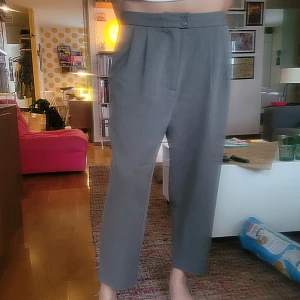 Grå kostymbyxor med hög midja - Säljer ett par grå kostymbyxor med hög midja och två knappar framtill. Byxorna har veckade detaljer vid midjan och raka Perfekta för en stilren och clean outfit. Fint begagnat skick, dock några noppar vid ena fickan. 