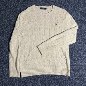 Grå kabelstickad tröja från Ralph Lauren - Säljer en klassisk beige kabelstickad tröja från Polo Ralph Lauren med rund halsringning och broderad logga på bröstet. Tröjan har långa ärmar och ribbade muddar vid ärmslut och nederkant. Perfekt för en stilren och tidlös look.