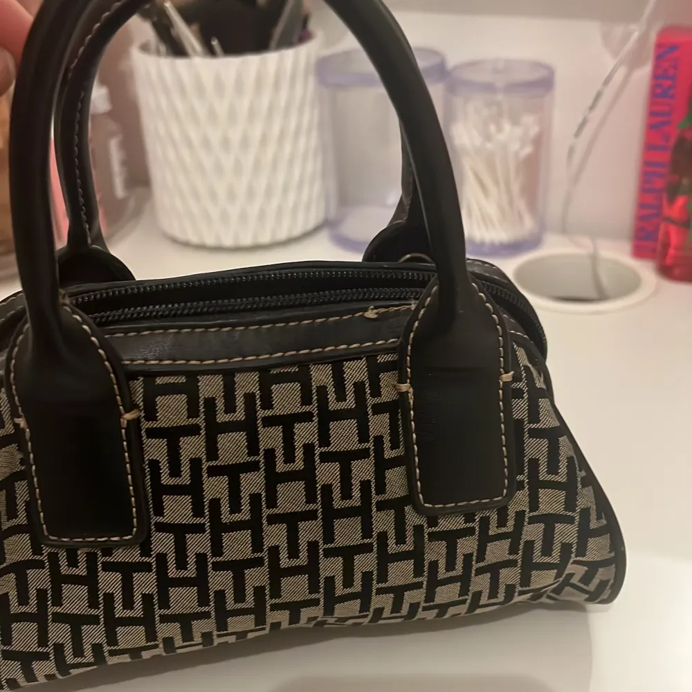 Säljer en snygg handväska från Tommy Hilfiger med svart och beige mönster, svarta handtag och detaljer i rött och grönt med guldfärgade knappar. Väskan har dragkedja och är perfekt för dig som vill ha en stilren accessoar.. Laukut & Käsilaukut.