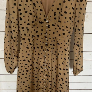 Adoore paris klänning - Säljer en beige långärmad klänning med svarta prickar och djup v-ringning. Klänningen har knappar längs hela framsidan och markerad midja. Perfekt för dig som gillar trendiga mönster och en feminin siluett.