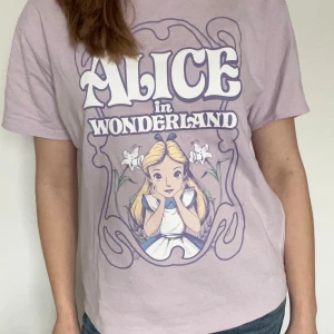 Ljuslila Alice i Underlandet t-shirt från H&M Divided - Säljer en ljuslila t-shirt från H&M Divided i storlek XS men som har en smått oversized passform. Den är använd endast ett fåtal gånger och är perfekt för dig som gillar Disney och vill ha en unik t-shirt!💜
