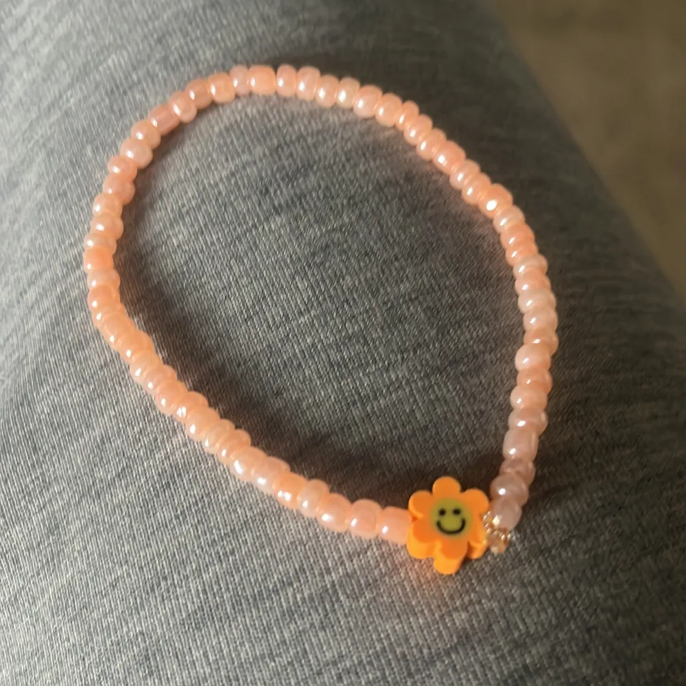 Sött armband med små rosa pärlor och en gul blomma med smiley. Perfekt accessoar för att liva upp din outfit med en lekfull detalj.. Asusteet.