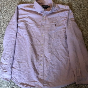 Ljuslila skjorta från Ralph Lauren - Klassisk ljuslila skjorta från Ralph Lauren med button-down krage och broderad logga på bröstet. Skjortan har lång ärm och knäppning framtill. Perfekt för en stilren och tidlös look. Storlek Xl, inga fläckar eller likande.