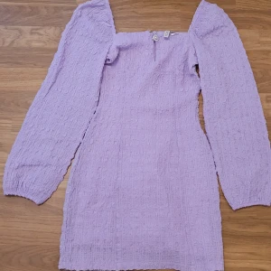 Ljuslila kort klänning med puffärmar - Säljer en superfin ljuslila kort klänning med puffiga långärmar och fyrkantig ringning. Klänningen har en strukturerad, ribbad känsla och markerad midja. Perfekt för dig som vill sticka ut med en trendig look!