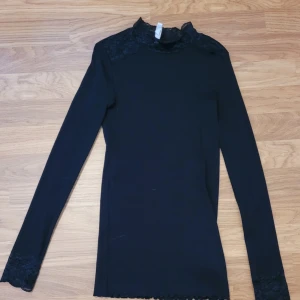 Svart långärmad topp med spetsdetaljer - Säljer en svart långärmad topp från Pomp de lux med fina spetsdetaljer vid halsringningen och axlarna. Perfekt att styla till olika outfits och ger en elegant touch med den höga spetskragen.