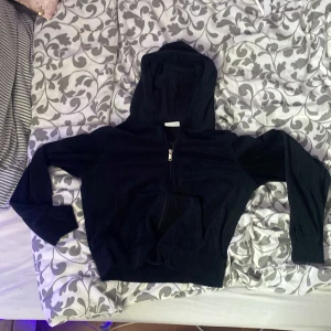 Svart hoodie med dragkedja från Kappahl - Säljer en svart hoodie från Kappahl med dragkedja framtill och huva. Tröjan har långa ärmar, ribbade muddar och en ficka framtill. Perfekt för en avslappnad stil.