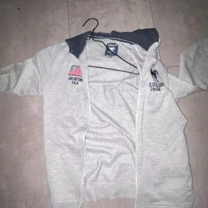 Ljusgrå zip-hoodie från Vinson Polo Club - Ljusgrå hoodie med dragkedja från Vinson Polo Club. Har broderad logga och amerikansk flagga på bröstet samt mörkblå detaljer i huvan. Perfekt för en avslappnad stil. Jag är 160 och den passar frf