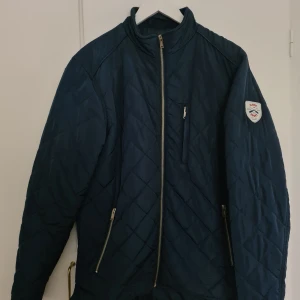 Mörkblå jacka från Lager 157 - Mörkblå quiltad jacka från Lager 157 med ståkrage och dragkedja framtill. Jackan har två sidofickor med dragkedja och en bröstficka. Märke på ärmen.