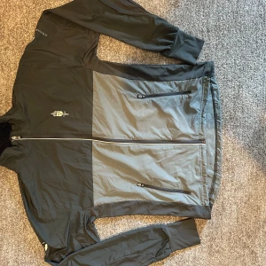 Trimtex tråningställ från förrsvarsmakten - Säljer en svart och grå vindjacka från Försvarsmakten med dragkedja framtill och två fickor med dragkedja. Jackan har lång ärm och tryckt logga på bröstet samt texten 'FÖRSVARSMAKTEN' på ryggen. Perfekt för träning utomhus. Storlek M