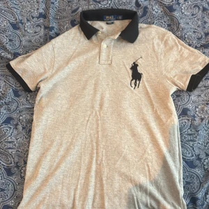 Grå pikétröja från Polo Ralph Lauren - Säljer en grå slim fit pikétröja från Polo Ralph Lauren med svart krage och ärmslut. Stor broderad logga på bröstet och stor text 'POLO' på baksidan av kragen. Klassisk och stilren design.