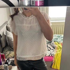 Vit blus med prickigt mönster och volang - Säljer en söt vit blus med små prickar och volangdetaljer vid ärmslut och nederkant. Blusen har korta ärmar och hög hals med spetskant. Perfekt för en fräsch och stilren look!