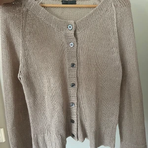 Beige stickad kofta med knappar - Säljer en beige stickad kofta med långa ärmar och rund halsringning. Koftan har synliga knappar framtill och ribbade muddar vid ärmslut och nederkant. Perfekt nu inför sommaren då den är väldigt tunn! Hör av er vid frågor 🥰